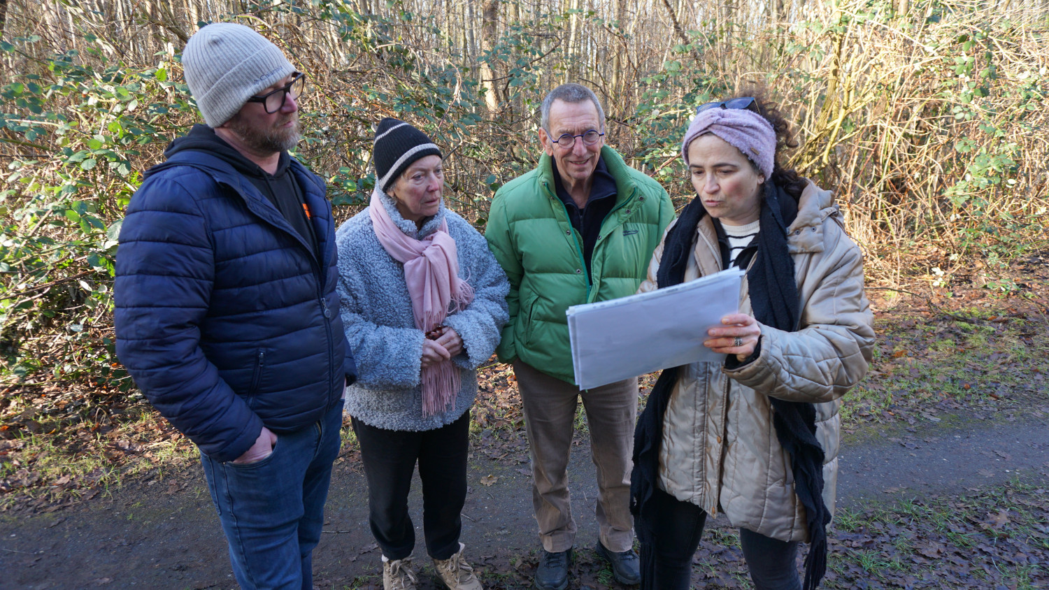Gezeigt werden vier Personen in der Natur. Rechts im Bild ist eine Frau mit Stirnband und Sonnenbrille auf dem Kopf, in der Hand hält sie in Klarsichtfolie eingepackte Notizzettel oder Bilder. Hierbei handelt es sich um Christel Trouvé, die wissenschaftliche Leitung des Denkort Bunker Valentin, die gerade die Familie von Fried Manders über das ehemalige Lagergelände führt. Links neben Christel steht ein Mann mit grüner Jacke und blauer Brille, neben ihm steht eine Frau mit flauschiger Jacke und rosafarbenem Schal, sie trägt dazu eine schwarze Mütze, und ganz links im Bild steht Goderd Manders mit Mütze und Brille sowie blauer Jacke, die Hände hat er in den Hosentaschen.  