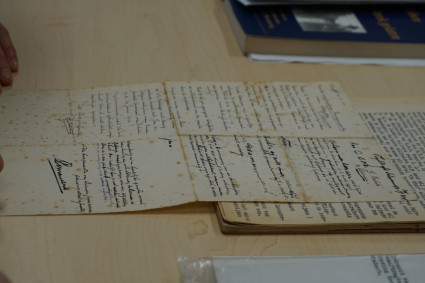 Links im Bild werden zwei Hände gezeigt, welche vom Licht ausgeblichene Notizblätter berühren. Auf dem Papier ist größtenteils in schwarz geschrieben worden, teilweise aber auch in blau. Im Hintergrund des Bildes befindet sich ein Ausschnitt eines dunkelblauen Buches. Im Vordergrund in Klarsichtfolie eingepackte Zettel, die aussehen, als seien sie mit einer Schreibmaschine beschrieben worden. 