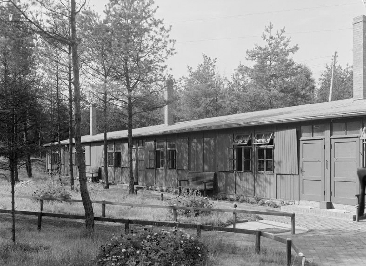 Abgebildet ist eine schwarz-weiß-Aufnahme einer Holzbaracke. Sie steht in der Mitte eines Waldgebietes. Im Vordergrund des Fotos sind niedrige, hölzerne Absperrungen für einen Gehweg zu sehen.  