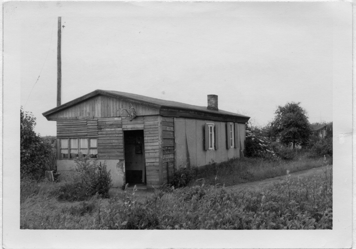 Gezeigt wird ein Foto einer Holzbaracke. Die Tür steht offen, das Gebäude ist weitläufig umgeben von einer Wiese und Gräsern. 