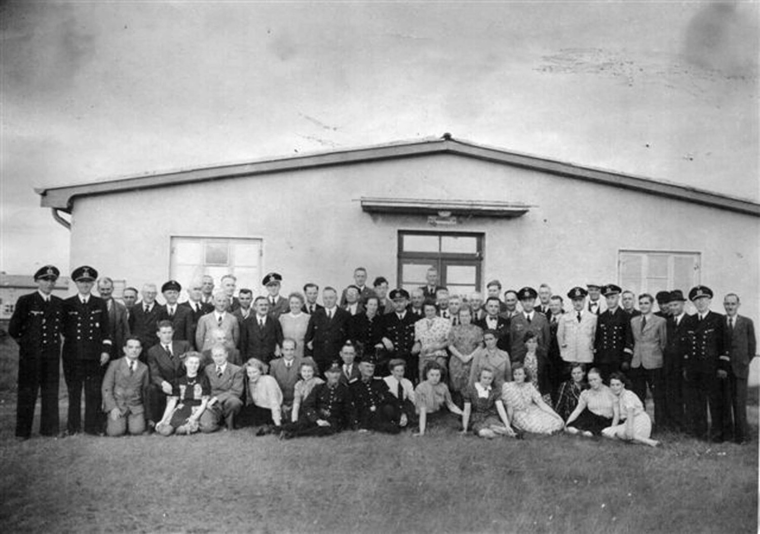 Abgebildet ist ein schwarz-weißes Gruppenfoto. Es zeigt uniformierte Männer sowie Frauen, die sehr schick gekleidet sind. Hinter der Menschenmenge ist die Baracke zu sehen.  
