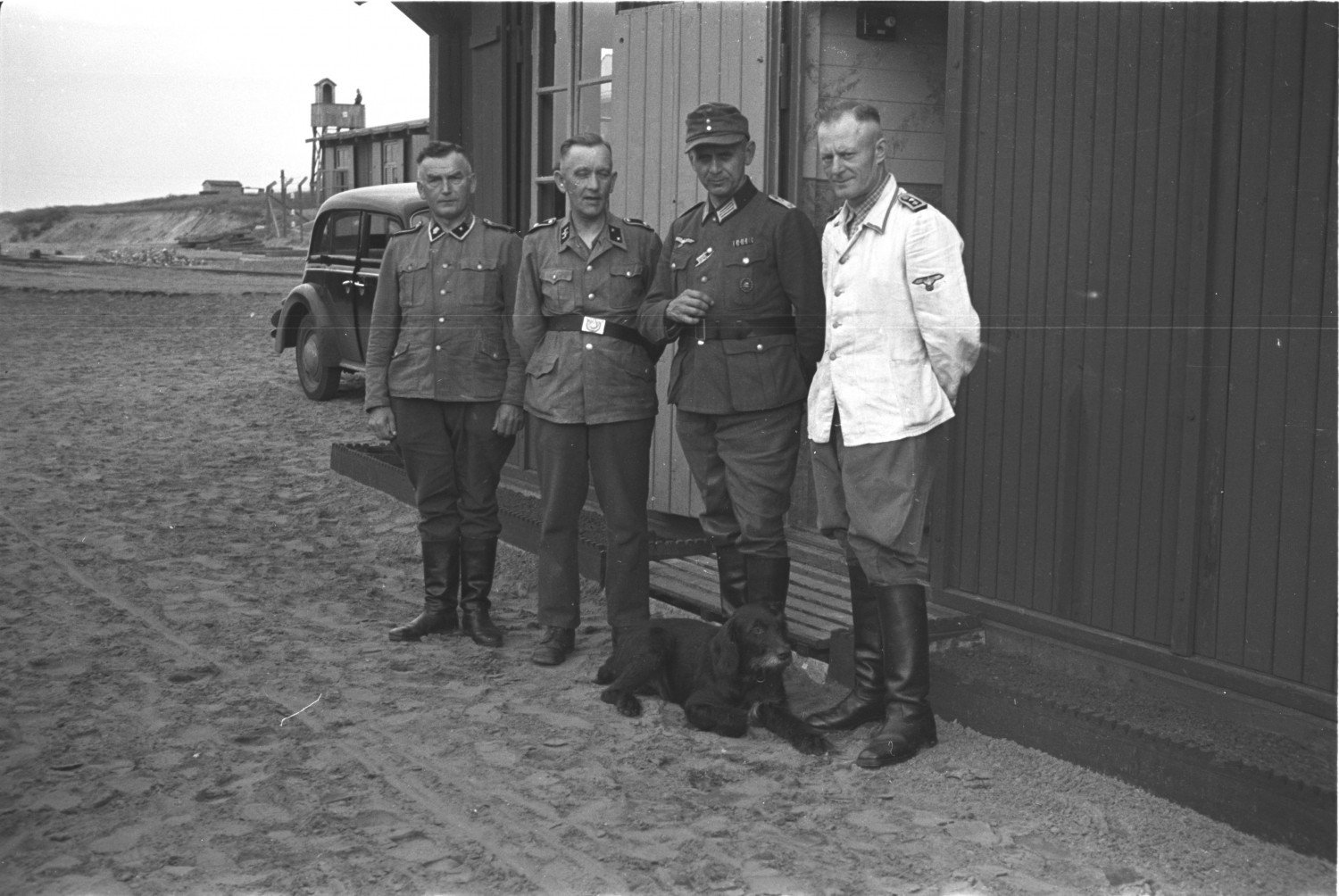 Abgebildet werden vier Männer und ein Hund. Hinter ihnen steht ein Gebäude, neben welchem ein Auto parkt. Sie stehen auf sandigem Untergrund. Alle sind uniformiert. Die Uniform des ganz rechts im Bild stehenden Mannes ist mit einer weißen Jacke versehen. 