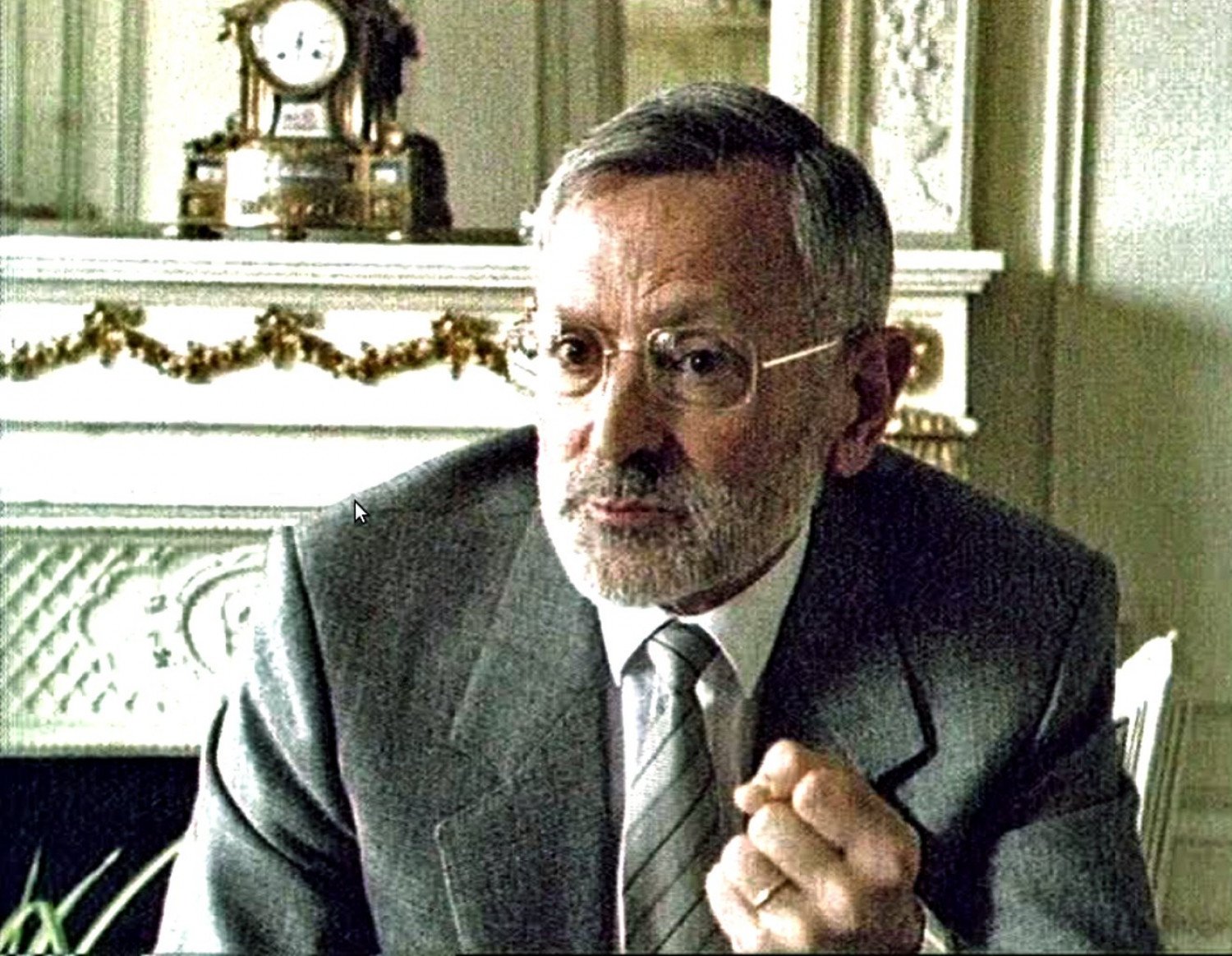 Gezeigt wird eine Momentaufnahme aus einem Interview, im Vordergrund sieht man einen <br />
Mann mit Brille und einem grauen Bart sowie grauem Haar. Er trägt einen Anzug mit Krawatte und Hemd und hat die linke Hand zur Faust geballt, weil er gerade spricht. Im Hintergrund sieht man eine Uhr und einen Ofen zum heizen.  