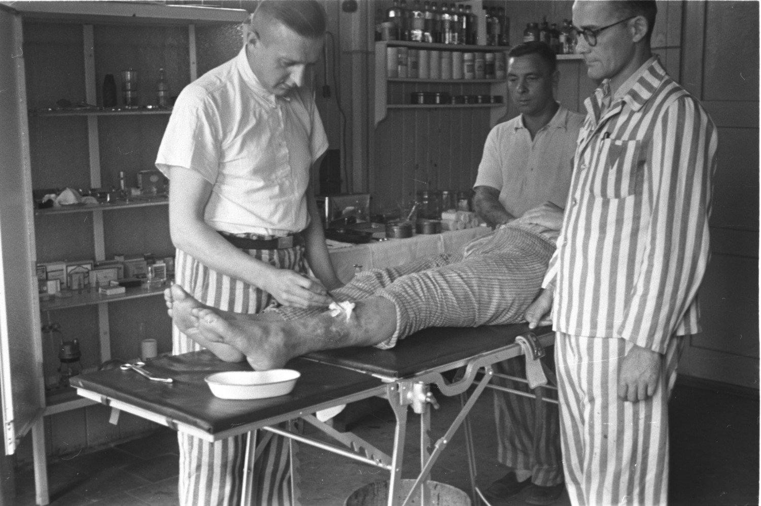 Gezeigt wird eine Aufnahme von vier Personen. Im rechten Vordergrund steht ein Mann mit einer gestreiften Hose, rechts im Bild steht ein Mann mit einem komplett gestreiften Outfit und im Hintergrund sieht man ebenfalls einen Mann mit gestreifter Hose. Sie stehen um eine Liege herum, auf welcher ein mutmaßlich verwundeter Mann liegt. Der links im Bild stehende Mann behandelt eine Wunde des Patienten am Schienbein. Die anderen Männer beobachten ihn dabei.  