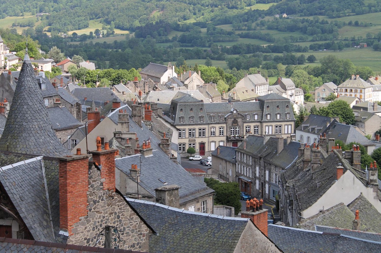 Sommerlicher Blick über Hausdächer auf einen kleinen Platz mit imposanten Gebäude, dahinter begrünte Landschaft.  