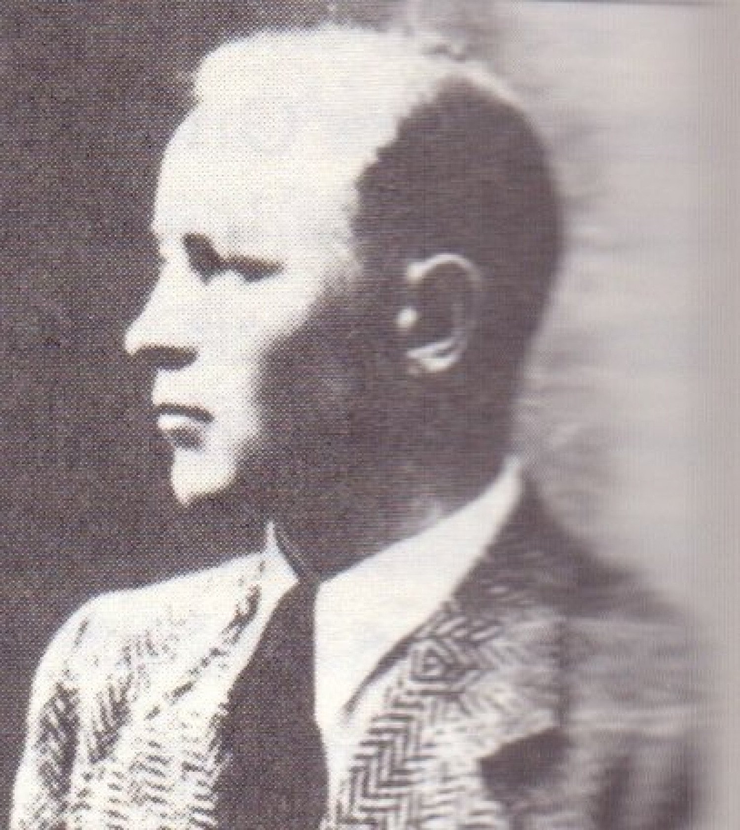 Gezeigt wird ein seitlich aufgenommenes Porträt in sepia-farben. Der Mann auf dem Bild trägt einen Anzug mit Hemd und Krawatte und blickt zur Seite, der Blick ist ernst. 