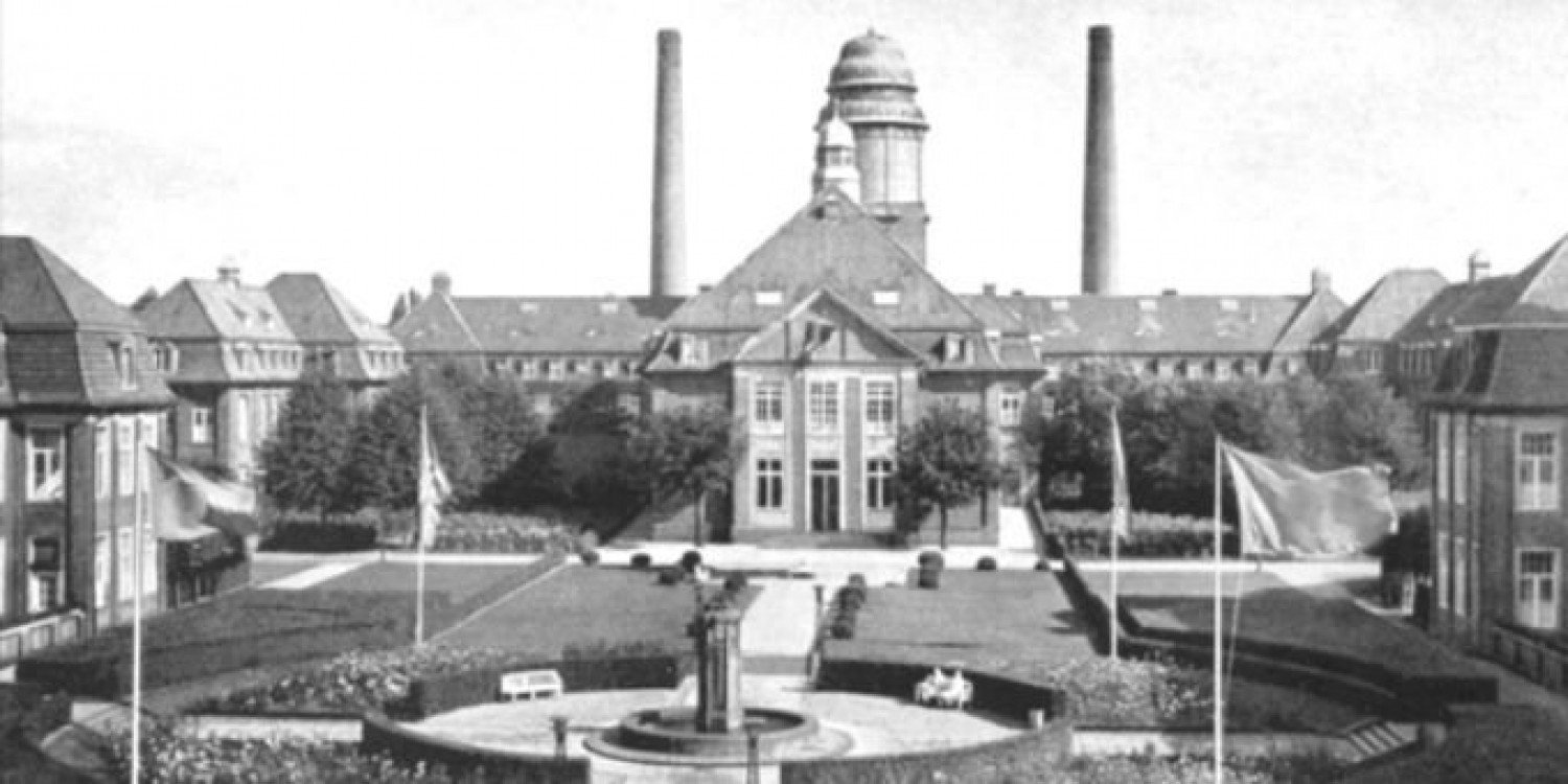 Gezeigt wird ein schwarz-weißes Bild, welches ein großes Gebäude abbildet und den sehr großen Garten, der sich vor dem Gebäude befindet. Im Vordergrund sind Fahnen zu sehen, die im Wind wehen. Mittig, im Garten, steht eine Art Springbrunnen innerhalb eines Kreises.  