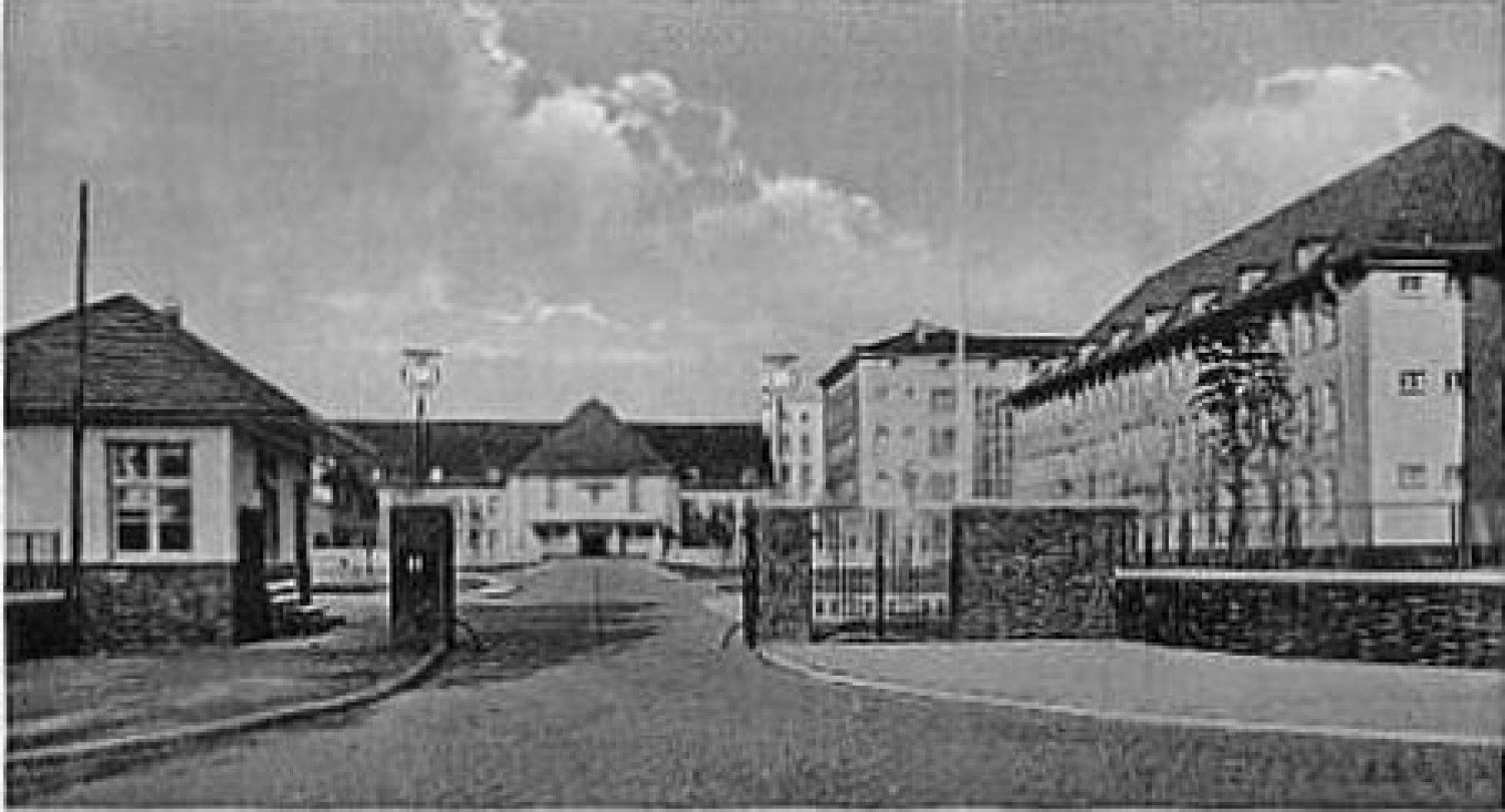 Abgebildet wird ein schwarz-weißes Foto von mehreren Häusern. Mittig ist ein Tor abgebildet, welches offen steht. Durch das Tor führt eine Straße. 