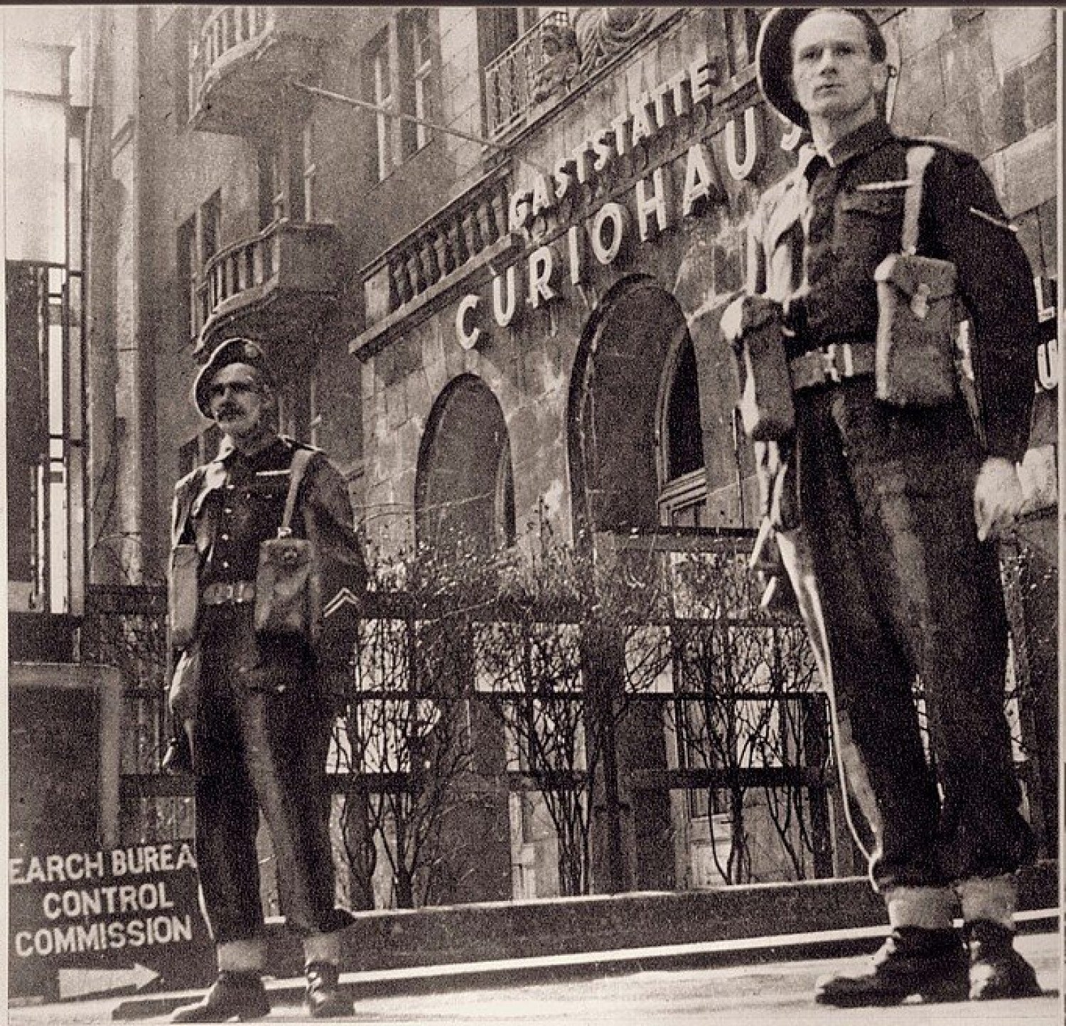 Es ist ein sepiafarbenes Bild abgebildet. Im Vordergrund stehen zwei Wachmänner, im Hintergrund ist die Gaststätte Curiohaus zu sehen. Im linken unteren Bildrand steht abgeschnitten: englisch "Search Bureau Control Commission" mit einem Pfeil nach rechts. Die beiden Männer schauen an der Kamera vorbei, der Blick ist sehr ernst. Vor dem Gebäude des Curiohauses sind kahle Sträucher zu sehen.  