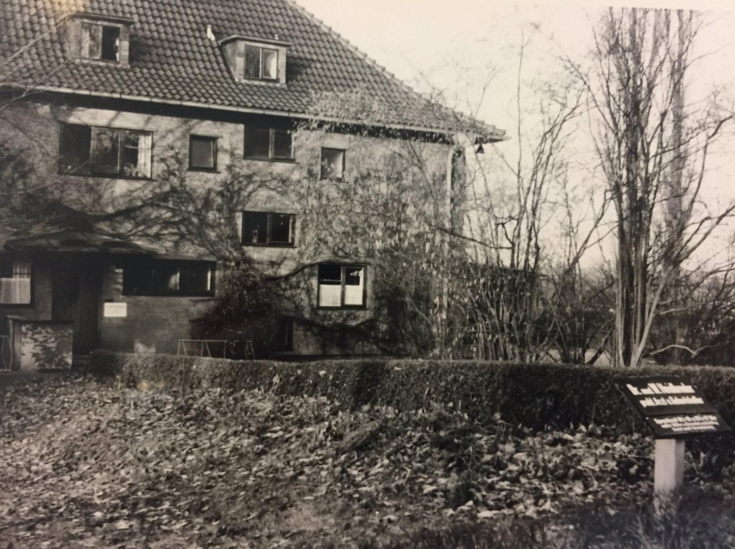 Gezeigt wird ein sepia-farbenes Bild von einem Haus, im Vordergrund wird die Hecke mitsamt dem Garten des Hauses abgebildet. In der rechten, vorderen Ecke steht ein Schild - die Inschrift ist nicht lesbar. 