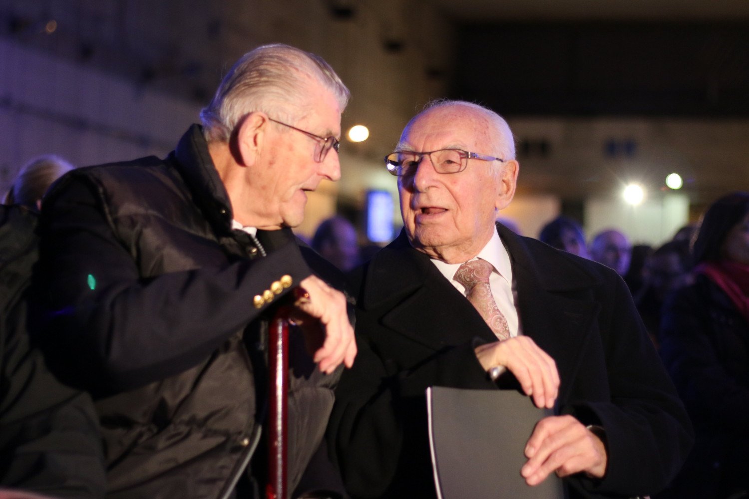 Abgebildet sind zwei ältere Männer, sie tragen eine Brille und haben weißes Haar. Die Aufnahme ist entstanden, während sich die beiden Männer unterhalten.  