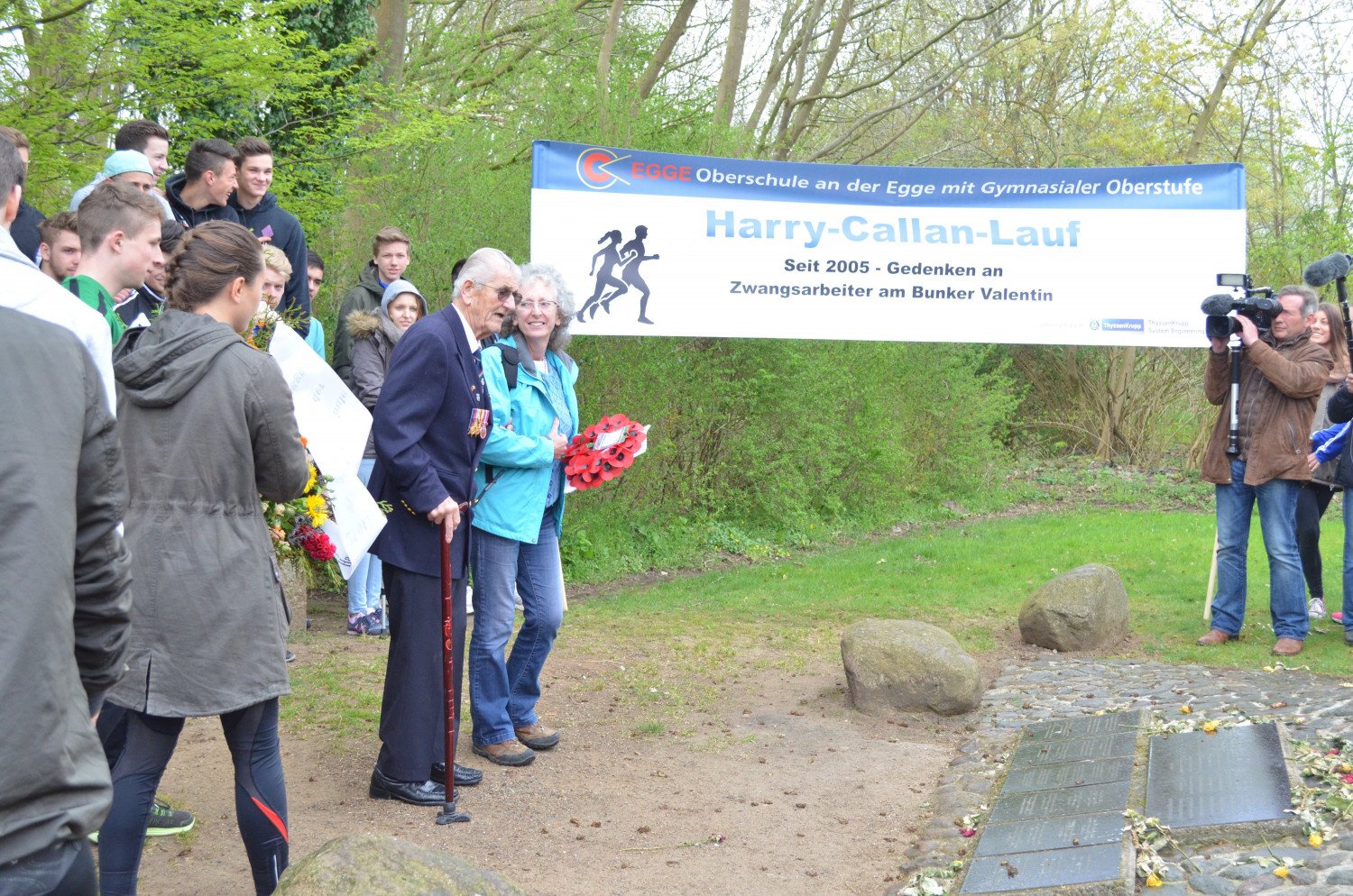 Abgebildet wird ein Foto von mehreren Personen. In der linken Hälfte des Bildes sieht man junge Menschen, die in Sportkleidung und mit dem Rücken zur Kamera gewandt stehen. Vor ihnen steht Harry Callan mit Gehstock und Anzug, neben ihm steht seine Schwiegertochter. Rechts im Bild sind Menschen zu sehen, die Kameras in der Hand halten. Mittig des Bildes ist ein Banner mit der Aufschrift "Harry-Callan-Lauf" zu sehen. 