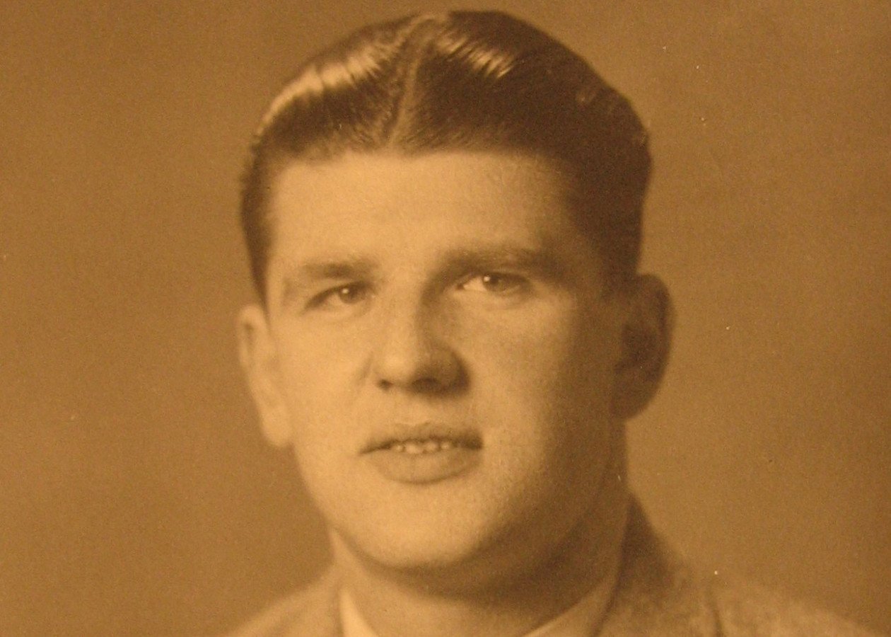 Gezeigt wird ein sepia-farbenes Bild von einem jungen Mann. Es ist eine Porträtaufnahme, der Mann trägt Anzug mit Hemd und Krawatte. Die Haare sind sehr glatt, er blickt direkt in die Kamera.