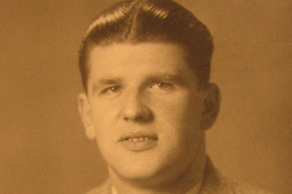 Gezeigt wird ein sepia-farbenes Bild von einem jungen Mann. Es ist eine Porträtaufnahme, der Mann trägt Anzug mit Hemd und Krawatte. Die Haare sind sehr glatt, er blickt direkt in die Kamera.