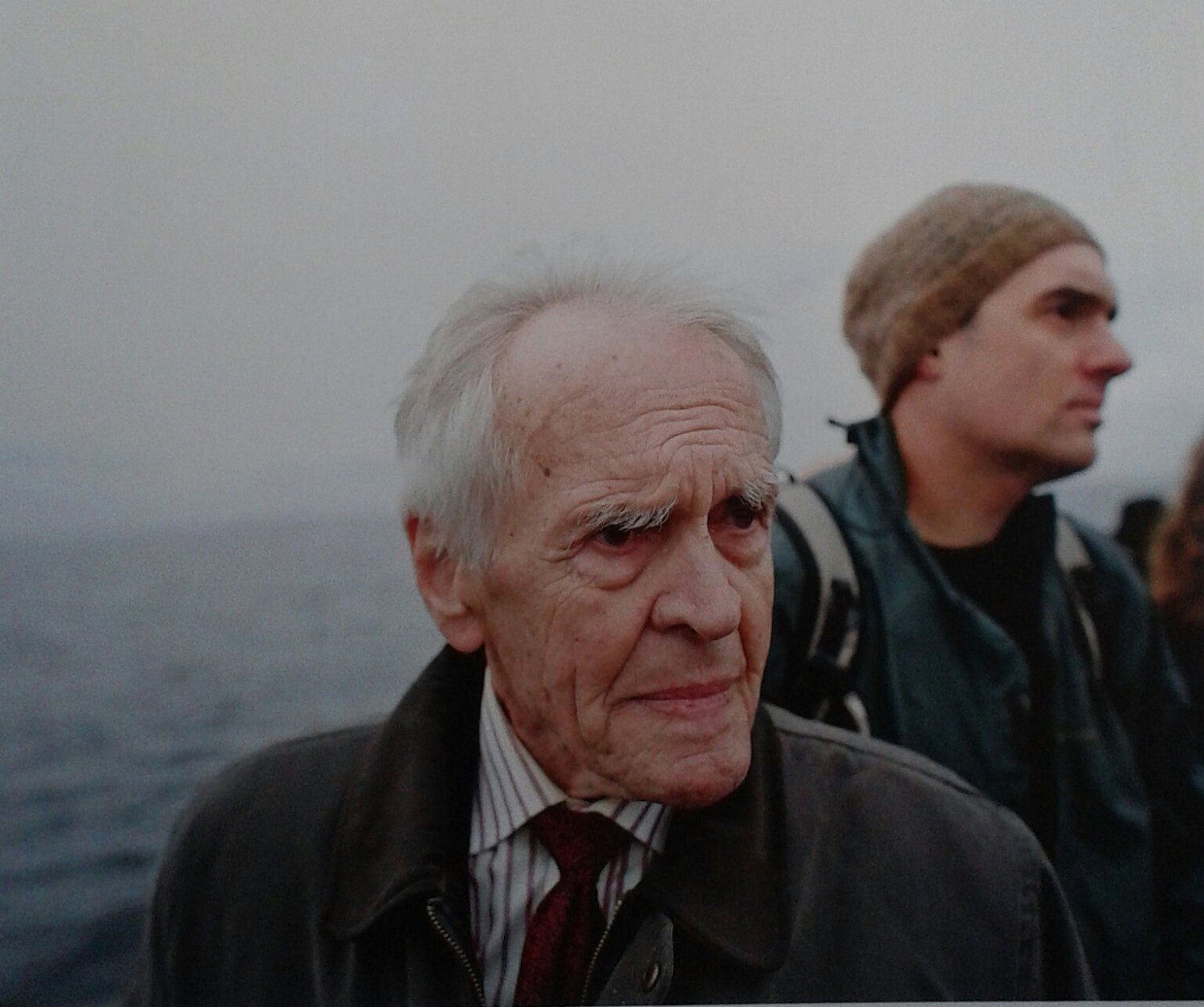 Ein älterer Mann blickt seitlich an der Kamera vorbei. Im Hintergrund ist das Meer zu sehen und ein jüngerer Mann mit einer Wollmütze. 