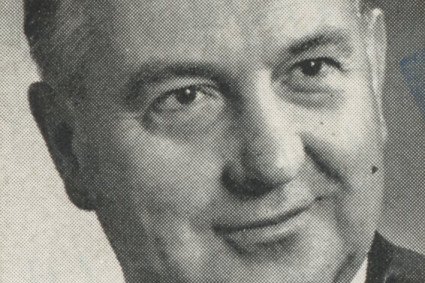 Abgebildet wird eine schwarz-weiße Nahaufnahme von einem älteren Mann. Er trägt ein Sakko mit Hemd und Krawatte und blickt verschmitzt grinsend in die Kamera.