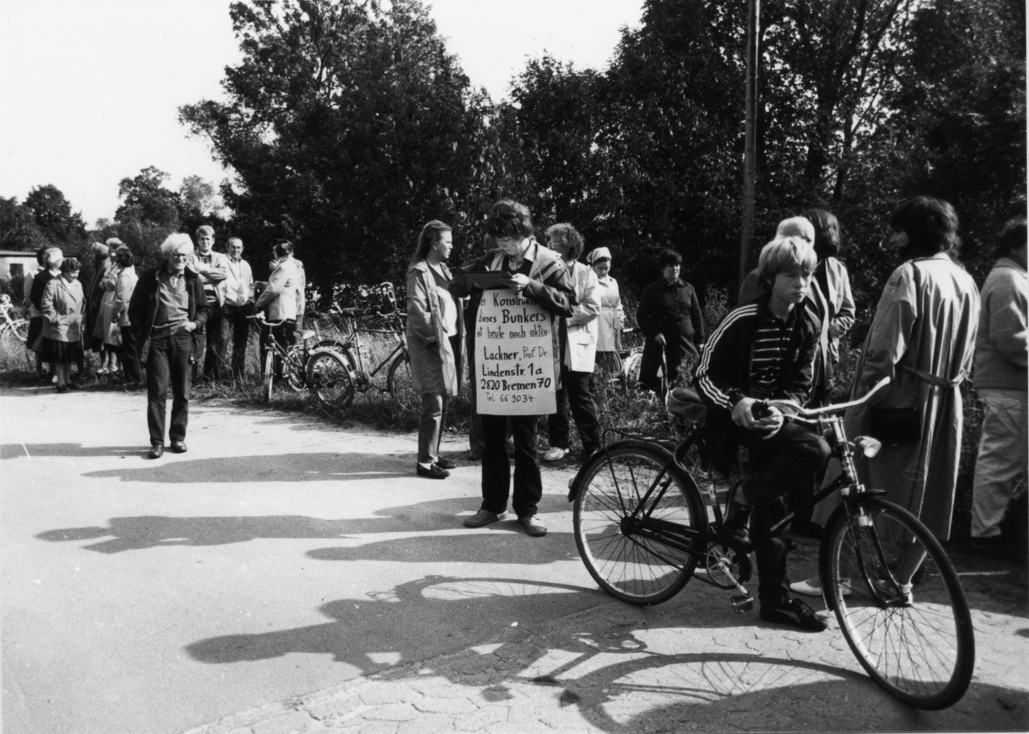 Gezeigt wird ein schwarz-weißes Bild. Es zeigt eine Ansammlung von Menschen. Im Vordergrund ist ein Junge auf einem Fahrrad zu sehen, mittig des Bildes steht eine Person mit einem Plakat. Im Hintergrund befinden sich weitere Menschen, teils mit Fahrrad. Weitere Informationen zum Bild finden Sie im Transkript des Bildes