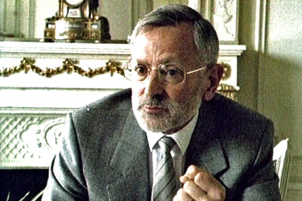 Gezeigt wird eine Momentaufnahme aus einem Interview, im Vordergrund sieht man einen <br />
Mann mit Brille und einem grauen Bart sowie grauem Haar. Er trägt einen Anzug mit Krawatte und Hemd und hat die linke Hand zur Faust geballt, weil er gerade spricht. Im Hintergrund sieht man eine Uhr und einen Ofen zum heizen. 