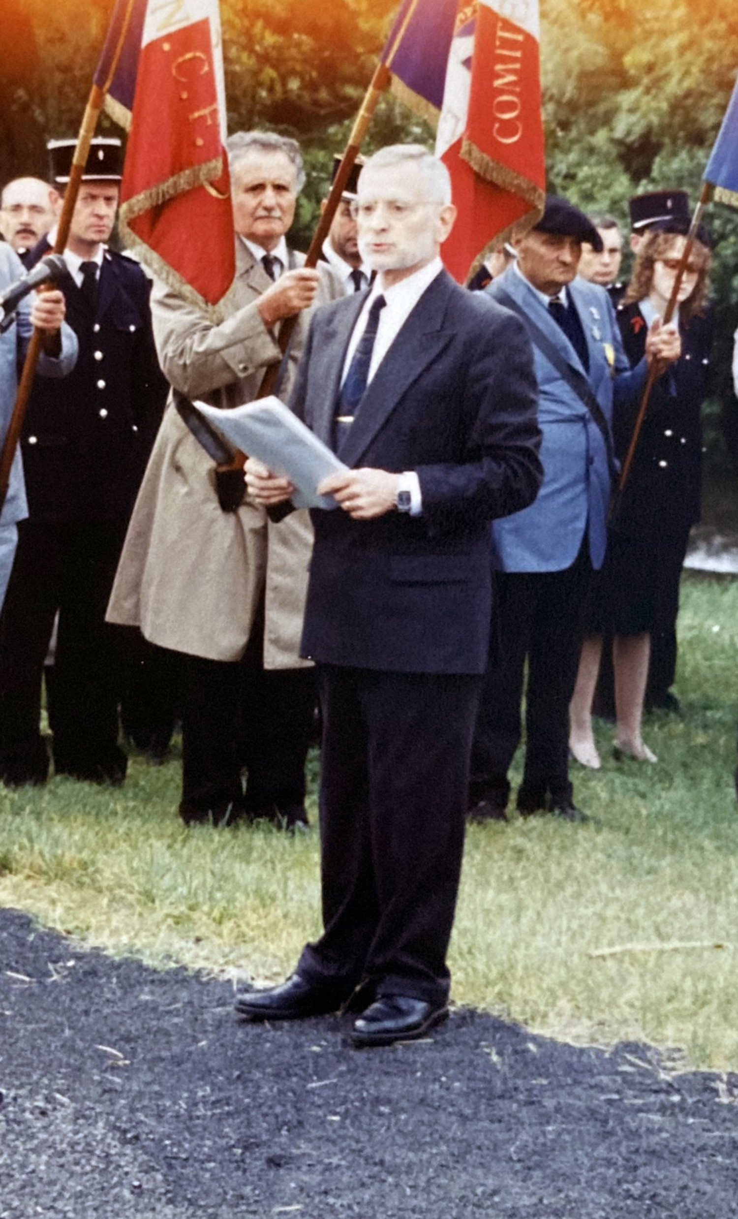 Gezeigt wird ein Foto, welches mehrere Menschen abbildet. Im Vordergrund ist ein älterer Mann mit Bart zu sehen, er trägt einen Anzug mit Krawatte und Hemd und hält in seinen Händen eine Mappe, aus der er vorliest. Hinter ihm stehen verschiedene Personen, die teilweise eine französische Flagge halten.  