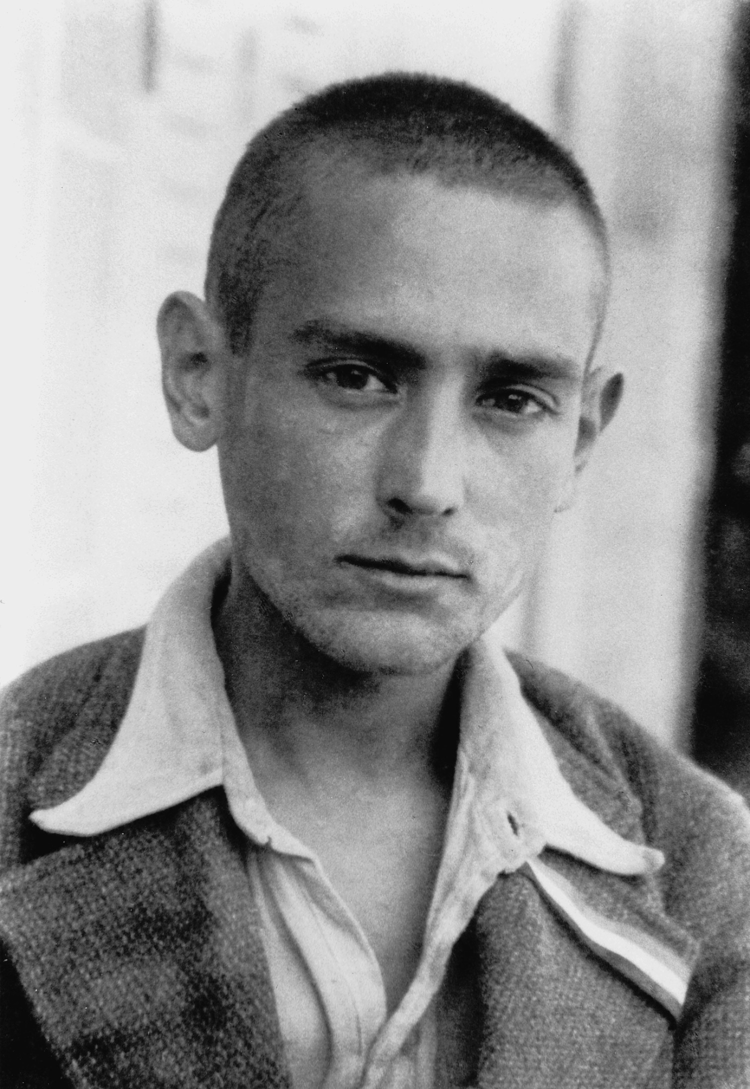 Gezeigt wird ein schwarz-weißes Foto, es ist eine Nahaufnahme von einer jungen, männlich gelesenen Person. Er hat kurz geschorene Haare und trägt ein Hemd, der oberste Knopf wurde offen gelassen. Dazu trägt er ein Sakko. Der Blick ist eher ernst, aber trotzdem lächelt der Mann leicht.  