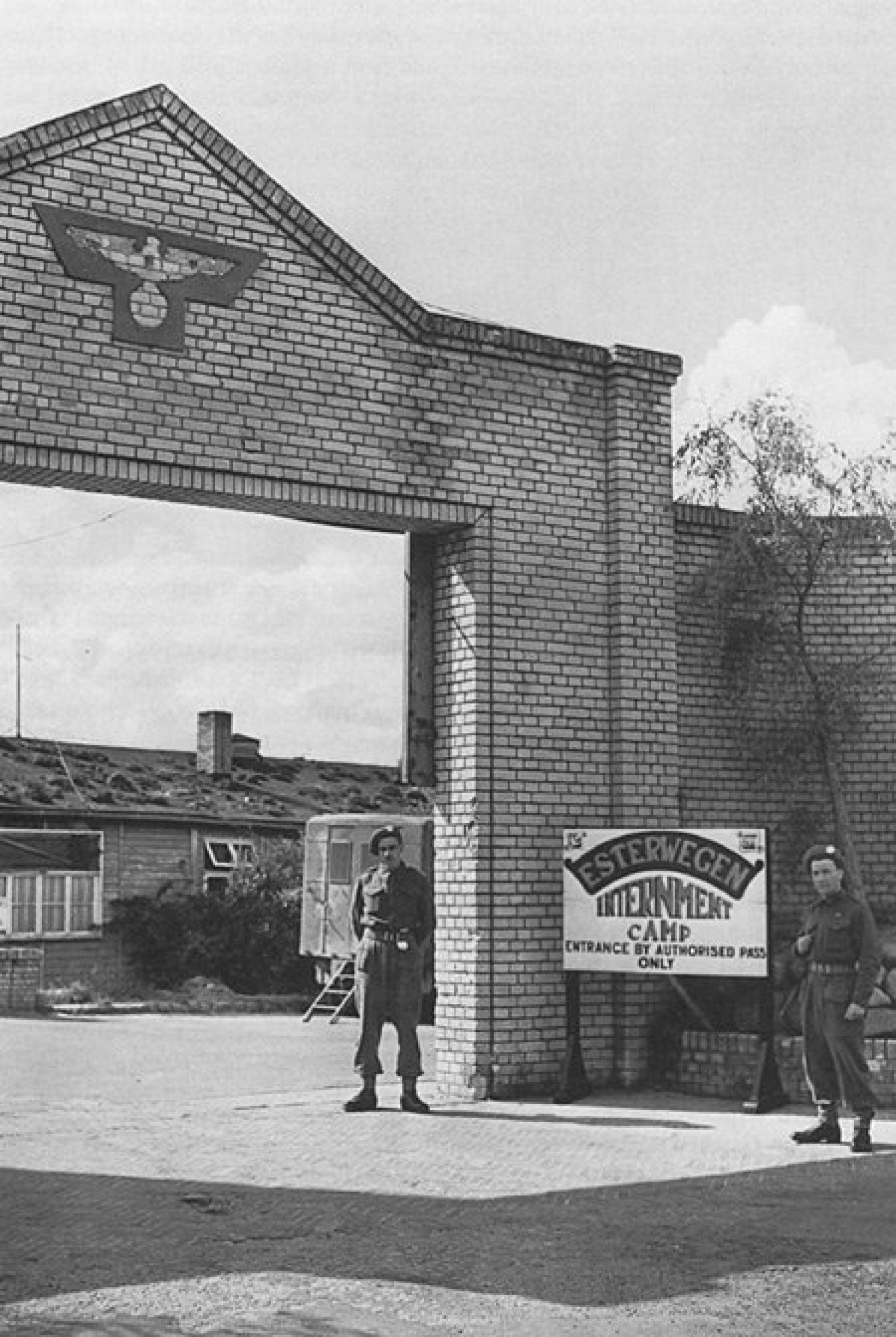 Abgebildet wird ein schwarz-weißes Foto, mittig ist ein gepflasterter Durchgang zu sehen, unter welchem ein Soldat steht. Rechts im Bild steht ein Schild mit der Inschrift: "Esterwegen internment camp", rechts neben dem Schild steht ebenfalls ein Soldat. 