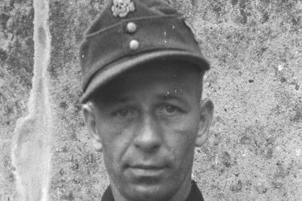 Gezeigt wird eine Nahaufnahme von einem Mann, das Foto wurde schwarz-weiß aufgenommen. Er trägt eine Kopfbedeckung und blickt sehr ernst in die Kamera.