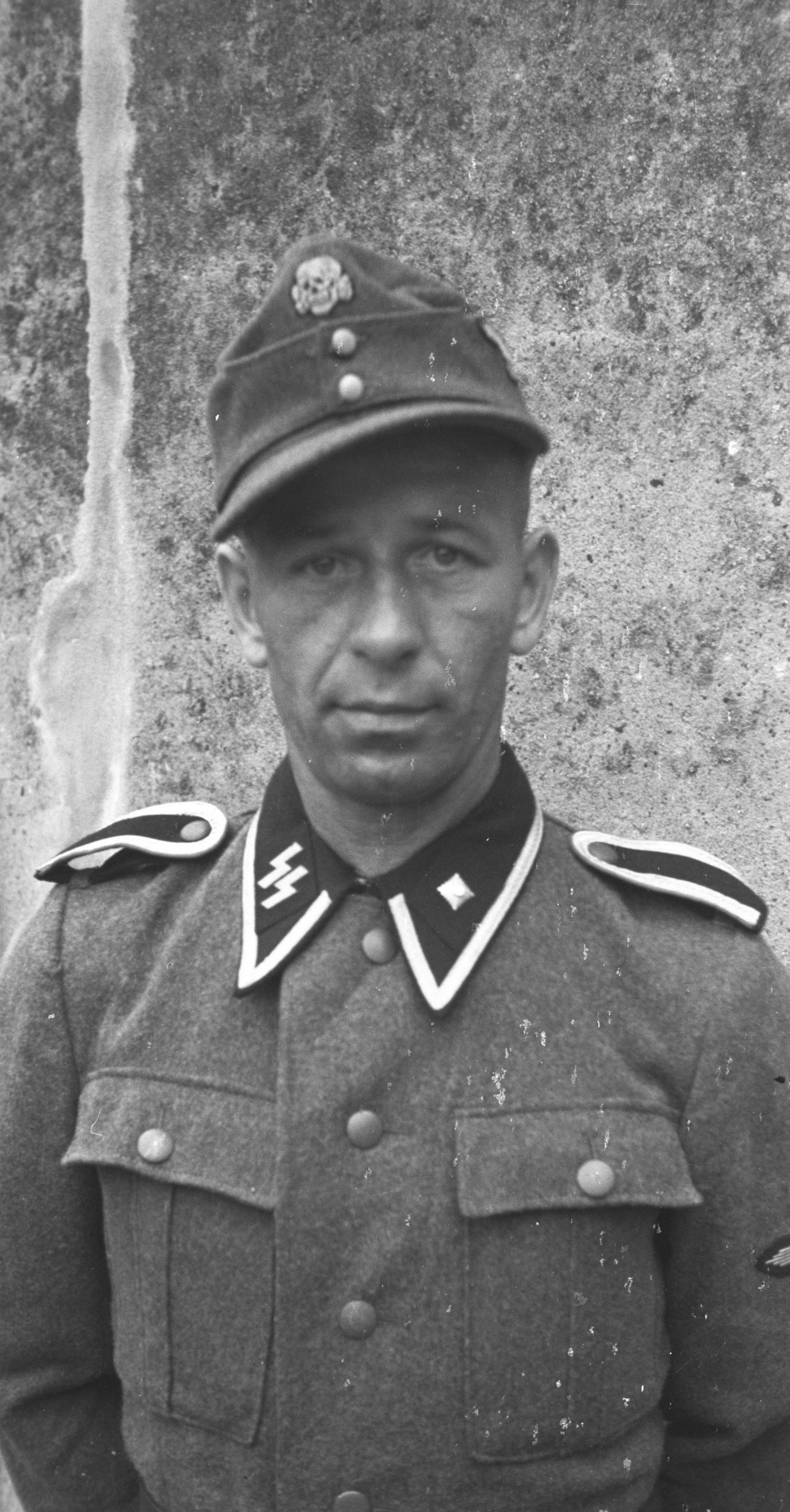 Gezeigt wird eine Nahaufnahme von einem Mann, das Foto wurde schwarz-weiß aufgenommen. Er trägt eine Kopfbedeckung und blickt sehr ernst in die Kamera. 