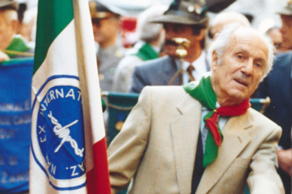 Elia Tomasi, auf diesem Foto schon älter, im Jahr 1994 bei einer Gedenk- oder Festveranstaltung, umgeben von weiteren Teilnehmenden. Er trägt ein Halstuch in den italienischen Farben und steht neben einer Fahne mit italienischer Trikolore. Er trägt die Fahne der Sektion Trient der ANEI (Nationaler Verband ehemaliger italienischer Militärinternierter)