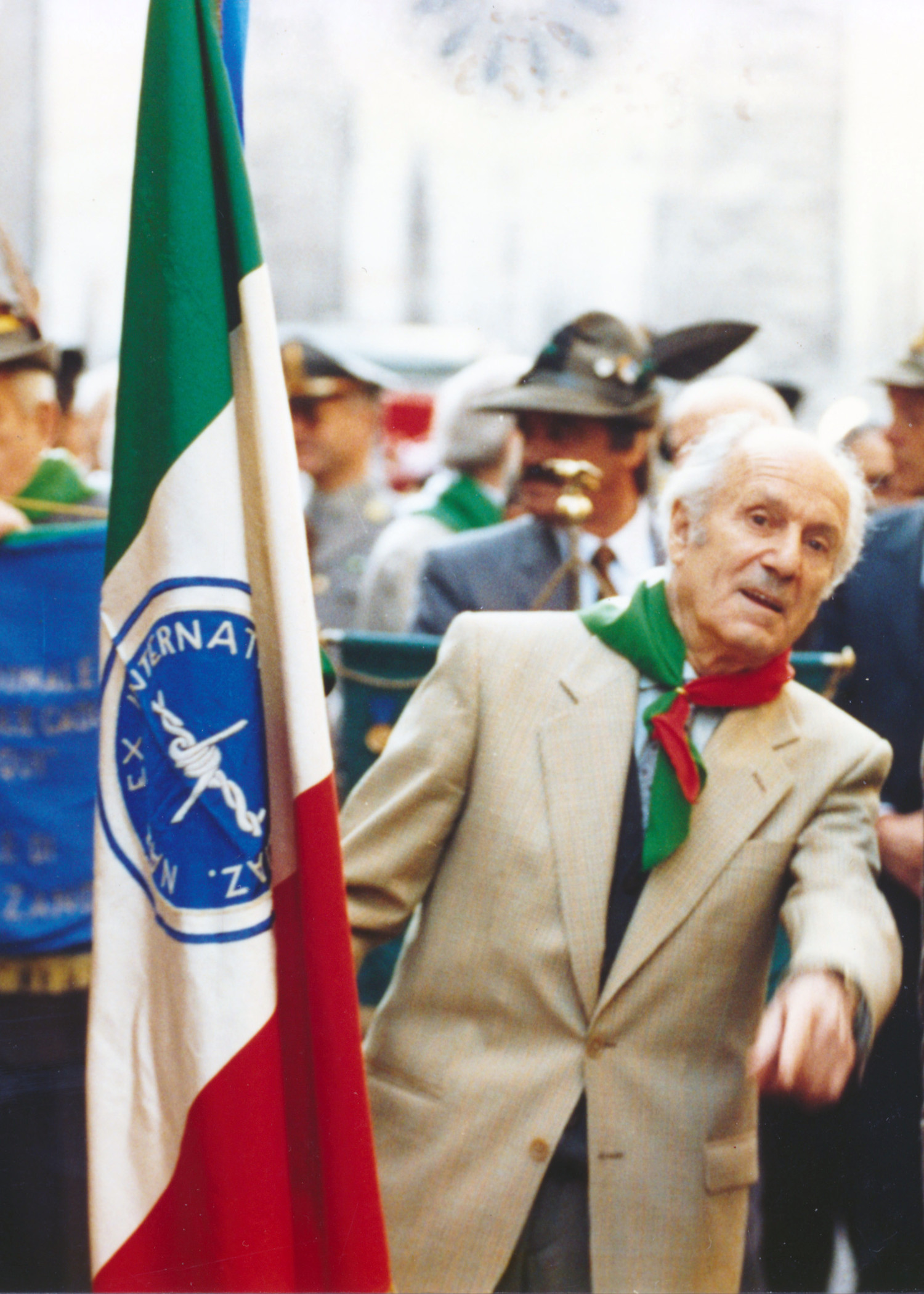 Elia Tomasi, auf diesem Foto schon älter, im Jahr 1994 bei einer Gedenk- oder Festveranstaltung, umgeben von weiteren Teilnehmenden. Er trägt ein Halstuch in den italienischen Farben und steht neben einer Fahne mit italienischer Trikolore. Er trägt die Fahne der Sektion Trient der ANEI (Nationaler Verband ehemaliger italienischer Militärinternierter) 