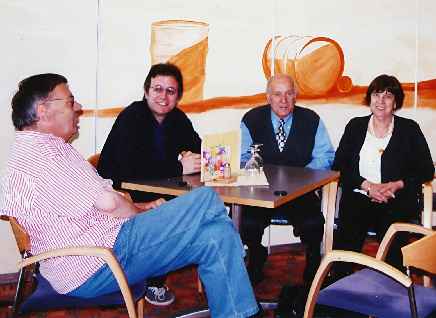 Das Foto zeigt vier Erwachsene, die gemeinsam an zwei zusammengestellten Tischen in einem hellen Raum sitzen. Die Gruppe sitzt in einem freundlich gestalteten Raum mit bemalten Wänden. Auf der Wand hinter ihnen ist ein abstraktes Stillleben zu sehen, das unter anderem ein großes Glas und eine umgefallene Tasse zeigt. Auf dem Tisch steht ein kleines dekoratives Arrangement mit bunten Bildern und zwei umgedrehten Weingläsern. Von rechts nach links: <br />
Person 1 sitzt ganz rechts. Hier handelt es sich um die Ehefrau von Elia, Carlina Tomasi. Sie trägt ein dunkles Jäckchen über einem weißen Oberteil und hat kurze, dunkle Haare. Auch diese Person lächelt leicht in Richtung Kamera.<br />
Person 2 sitzt rechts neben Person 1. Es handelt sich hier um Elia Tomasi. Er trägt ein hellblaues Hemd, eine dunkle Weste und eine gemusterte Krawatte. Elia Tomasi schaut freundlich in die Kamera.<br />
Person 3 sitzt am Tisch gegenüber der vierten Person, hier handelt es sich um Rainer Habel. Er trägt eine dunkle Jacke und eine Brille. Er lächelt und hat die Hände locker auf dem Tisch gefaltet.<br />
Person 4  sitzt seitlich auf einem Holzstuhl mit blauem Sitzpolster. Hier handelt es sich um Elia Tomasis Sohn, Maurizio. Er trägt ein kurzärmliges Hemd mit roten und weißen Streifen sowie eine blaue Jeans. Die Beine sind übereinandergeschlagen, und die Person schaut nach rechts zu den anderen am Tisch.<br />
<br />
<br />
<br />
 