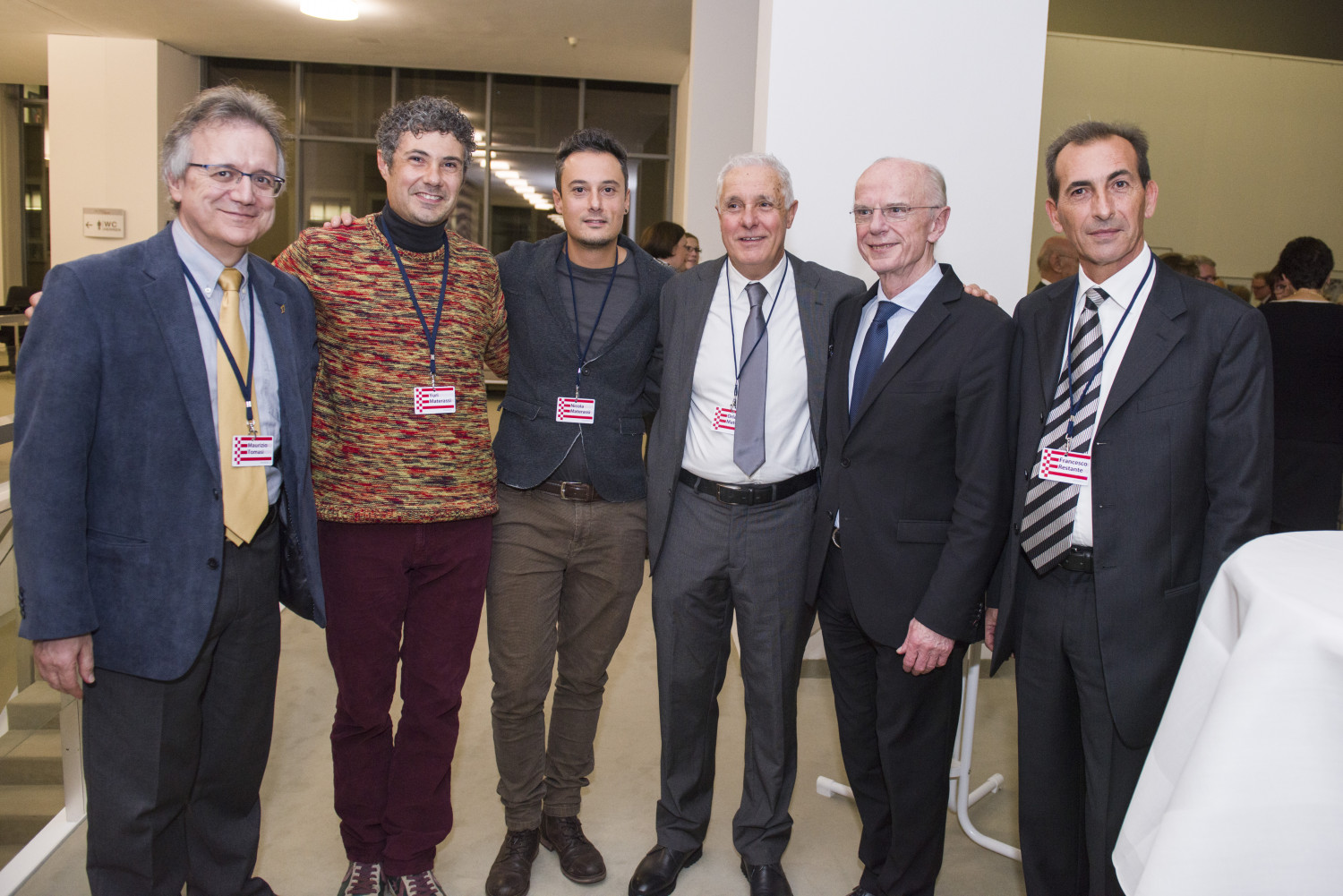 Das Foto zeigt sechs erwachsene Männer, die eng nebeneinander stehen und in die Kamera lächeln. Alle tragen sichtbar Namensschilder an blauen Bändern um den Hals.<br />
Ganz links steht ein Mann mit grauem Haar und Brille. Er trägt ein blaues Sakko, ein hellgelbes Hemd und eine gelbe Krawatte. Daneben steht ein Mann mit kurzen, gelockten dunklen Haaren. Er trägt einen mehrfarbig melierten Pullover in Rot- und Gelbtönen sowie eine weinrote Hose. Der dritte von links ist ein Mann mit kurzen dunklen Haaren und Drei-Tage-Bart. Er trägt ein dunkles Jackett, einen grauen Pullover und eine braune Hose. Er hat seine Arme locker um die beiden Männer neben ihm gelegt. Der vierte Mann hat kurzes graues Haar und trägt einen grauen Anzug mit weißem Hemd und grauer Krawatte. Er lächelt in die Kamera. Der fünfte Mann hat sehr kurz geschnittenes helles Haar und trägt einen dunklen Anzug mit weißem Hemd und dunkelblauer Krawatte. Er steht sehr aufrecht und wirkt formell gekleidet. Ganz rechts steht ein Mann mit kurzem dunklem Haar. Er trägt einen dunklen Anzug, ein weißes Hemd und eine gestreifte Krawatte. 