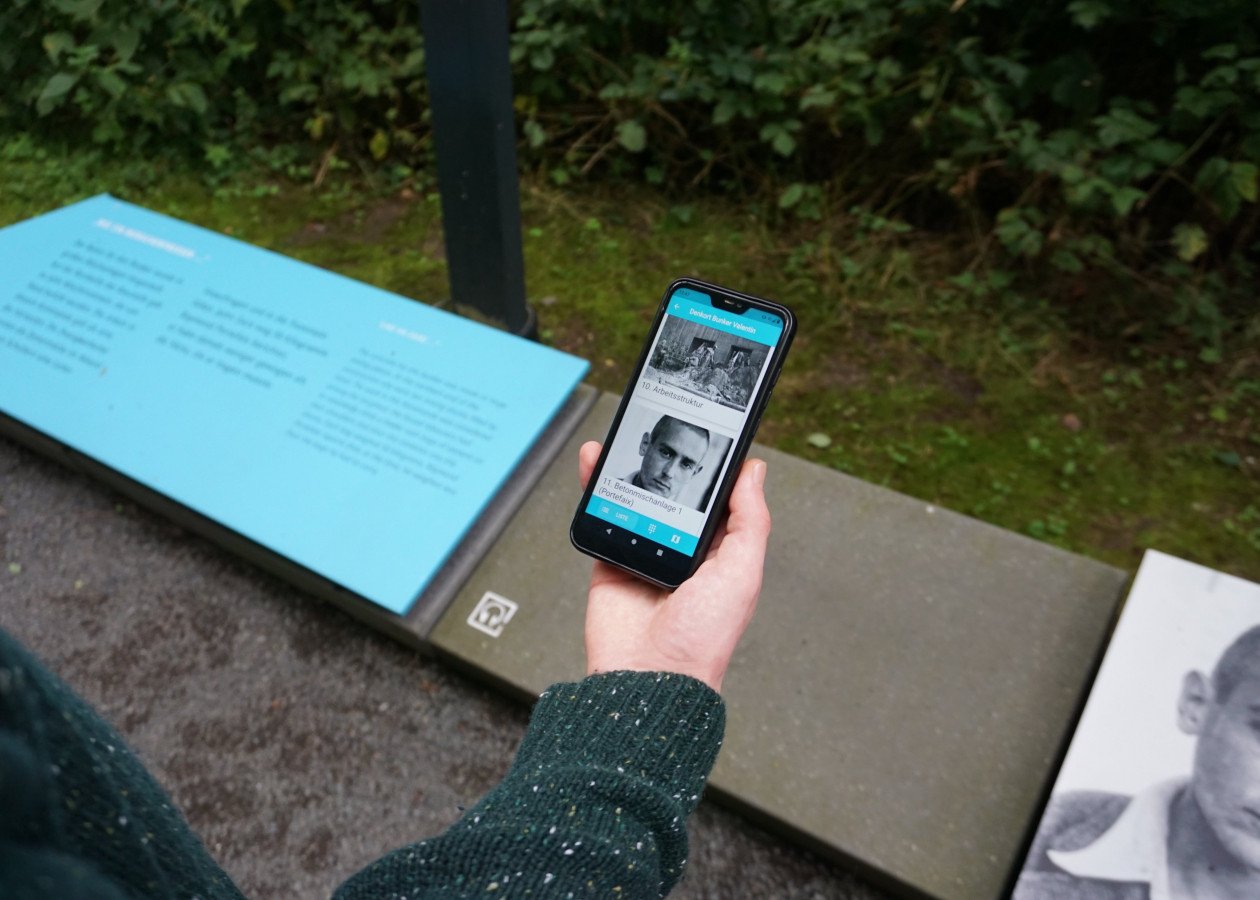 Ein Besucher steht an einer Station auf dem Rundweg und nutzt die App "Denkort Bunker Valentin" auf seinem Smartphone. Das Display zeigt die Einträge zu den Stationen "10. Arbeitsstruktur" und "11. Betonmischanlage 1 (Portefaix)". Ein Foto von Raymond Portefaix ist auf dem Display und auf den Infotafeln der Station zu sehen.