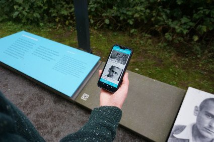 Ein Besucher steht an einer Station auf dem Rundweg und nutzt die App "Denkort Bunker Valentin" auf seinem Smartphone. Das Display zeigt die Einträge zu den Stationen "10. Arbeitsstruktur" und "11. Betonmischanlage 1 (Portefaix)". Ein Foto von Raymond Portefaix ist auf dem Display und auf den Infotafeln der Station zu sehen.