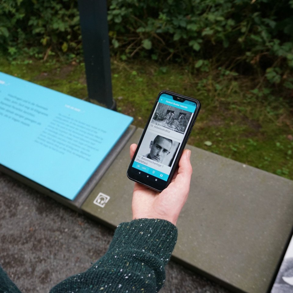 Ein Besucher steht an einer Station auf dem Rundweg und nutzt die App "Denkort Bunker Valentin" auf seinem Smartphone. Das Display zeigt die Einträge zu den Stationen "10. Arbeitsstruktur" und "11. Betonmischanlage 1 (Portefaix)". Ein Foto von Raymond Portefaix ist auf dem Display und auf den Infotafeln der Station zu sehen.