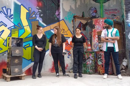 Vier jüngere Menschen stehen vor einer Wand, die mit bunten Graffitis bemalt ist. Links von ihnen steht ein Kunstwerk aus vier gestapelten Kartons. 