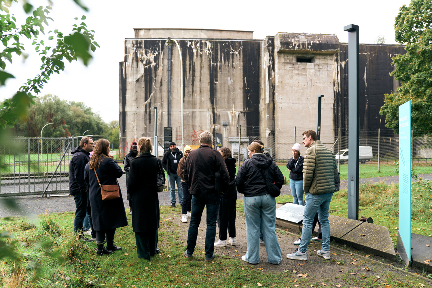 Auf dem Bild steht eine Gruppe von Erwachsenen im Freien vor dem massiven Betonbau des Bunker Valentin. Die Personen stehen in einem lockeren Kreis und hören offenbar einer Person zu, die etwas erklärt. Im Hintergrund ist der hohe, graue Bunker mit sichtbaren Altersspuren zu sehen, umgeben von Grünflächen und Bäumen. Rechts im Bild stehen Informationstafeln des Denkortes, links begrenzt ein Zaun das Gelände. Die Szene vermittelt den Eindruck eines geführten Austauschs oder Rundgangs am historischen Ort. 
