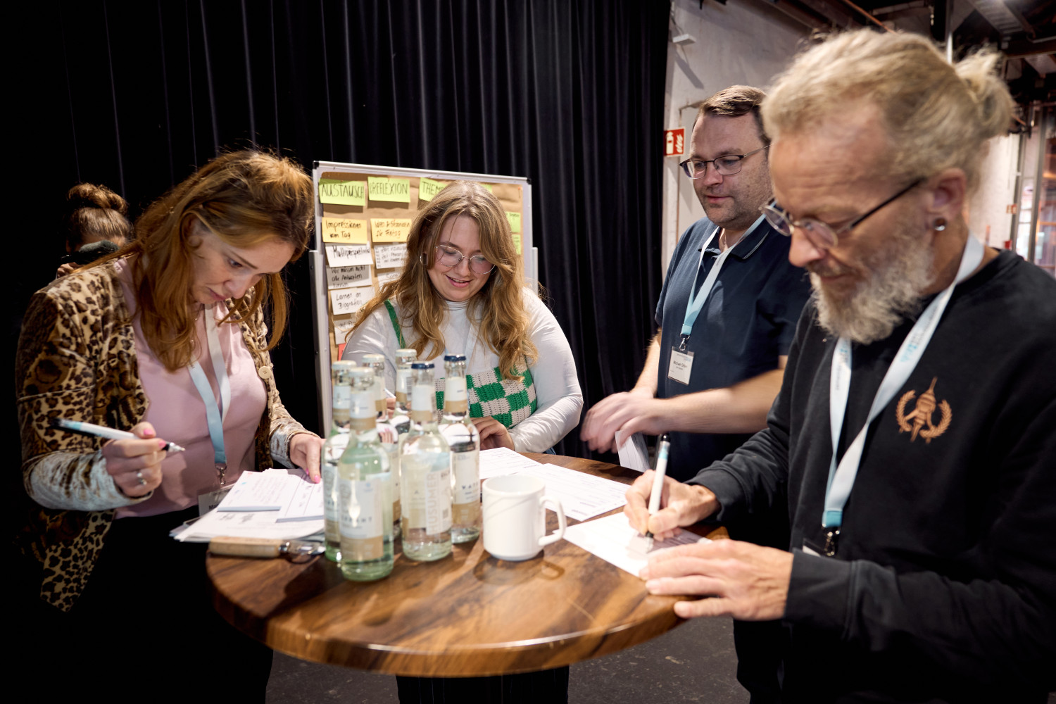 Auf dem Bild steht eine Gruppe von Erwachsenen an einem runden Stehtisch. Die Personen sind in eine gemeinsame Aufgabe vertieft und schreiben ihre Ideen auf Papier, das auf dem Tisch ausgebreitet liegt. Auf dem Tisch stehen zudem mehrere Wasserflaschen und eine Tasse. Alle tragen Namensschilder und scheinen miteinander im Austausch zu sein. Im Hintergrund ist eine Pinnwand mit Zetteln und Stichworten zu erkennen.  