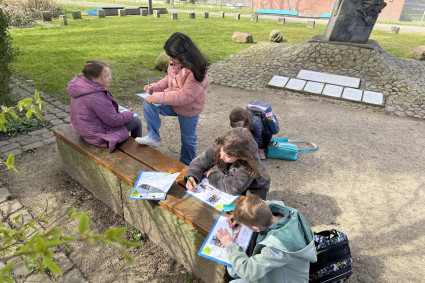 Eine Gruppe von Kindern im Freien, die an einer Bank sitzen und mit Heften arbeiten. Im Hintergrund ein Mahnmal. 