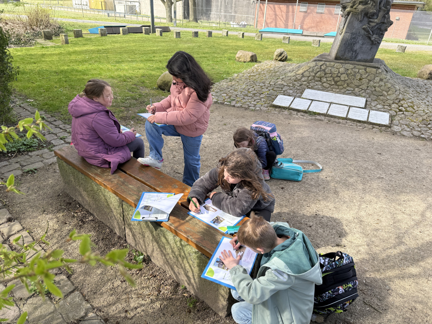 Eine Gruppe von Kindern im Freien, die an einer Bank sitzen und mit Heften arbeiten. Im Hintergrund ein Mahnmal.  
