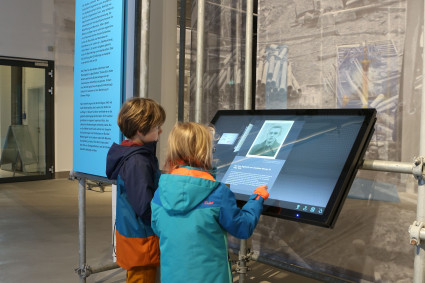 Zwei Kinder stehen in einem Ausstellungsraum vor einem großen, leicht schräg angebrachten Touchscreen. Beide tragen wetterfeste Jacken in Blau- und Orangetönen. Auf dem Bildschirm ist ein historisches Schwarz-Weiß-Foto einer Person sowie Text zu sehen. Links daneben steht eine hohe, hellblaue Infotafel mit der Überschrift „Der Blick der Verantwortlichen“. Im Hintergrund hängt ein großformatiges, graues Wandbild mit historischen Aufnahmen von Baustellen und Schienen. Die Umgebung wirkt modern und hell, mit Betonboden und Metallrohr-Konstruktionen, an denen die Ausstellungselemente befestigt sind.