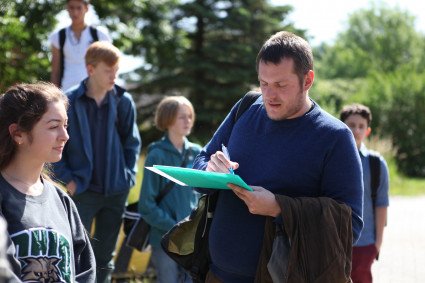 Ein Lehrer hält Zettel und Stift. Um ihn herum stehen Schüler:innen.