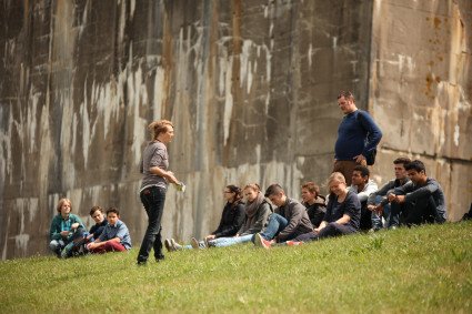 Eine Schulklasse sitzt auf dem Deich am Denkort Bunker Valentin. Ein Lehrer steht hinter der Gruppe. Die Seminarleiterin steht vor der Gruppe und hält Zettel in der Hand.