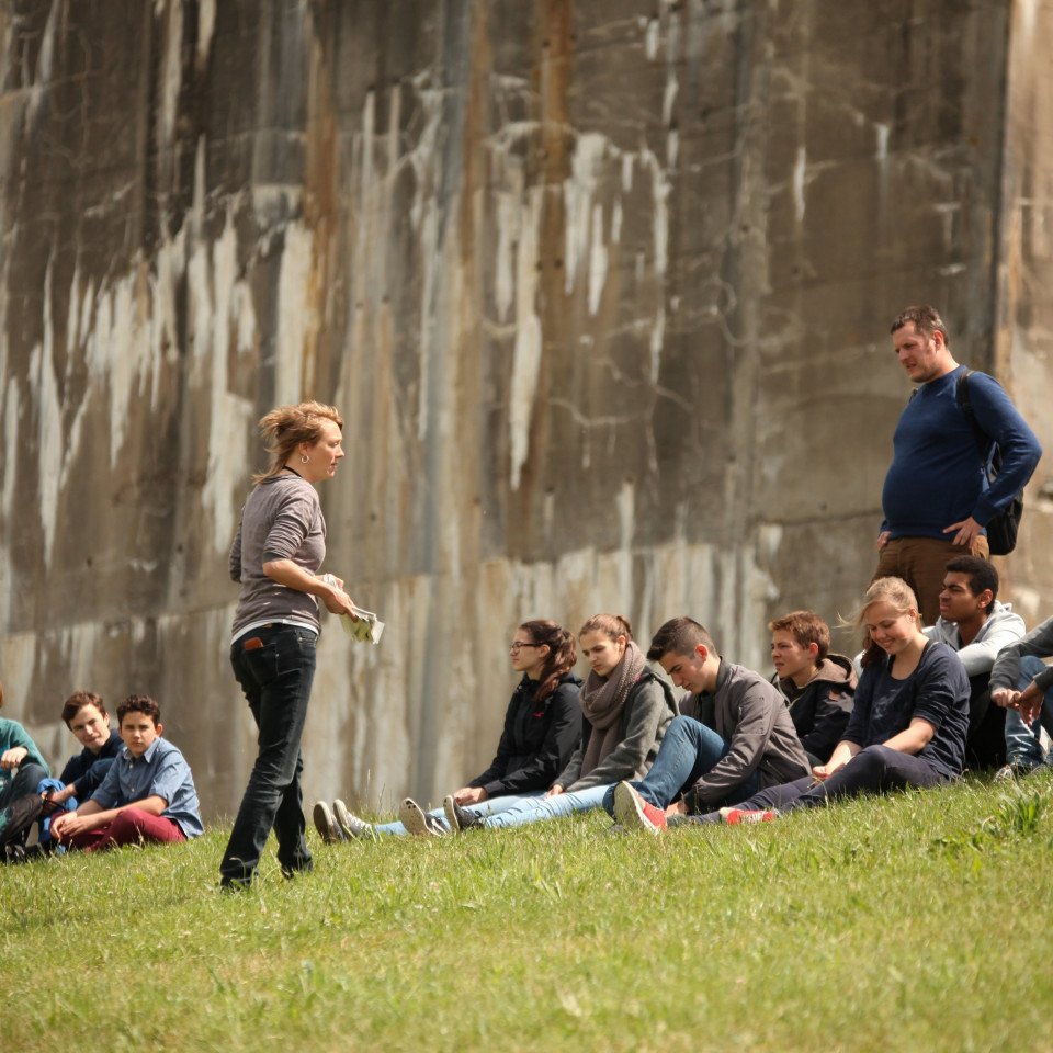 Eine Schulklasse sitzt auf dem Deich am Denkort Bunker Valentin. Ein Lehrer steht hinter der Gruppe. Die Seminarleiterin steht vor der Gruppe und hält Zettel in der Hand.