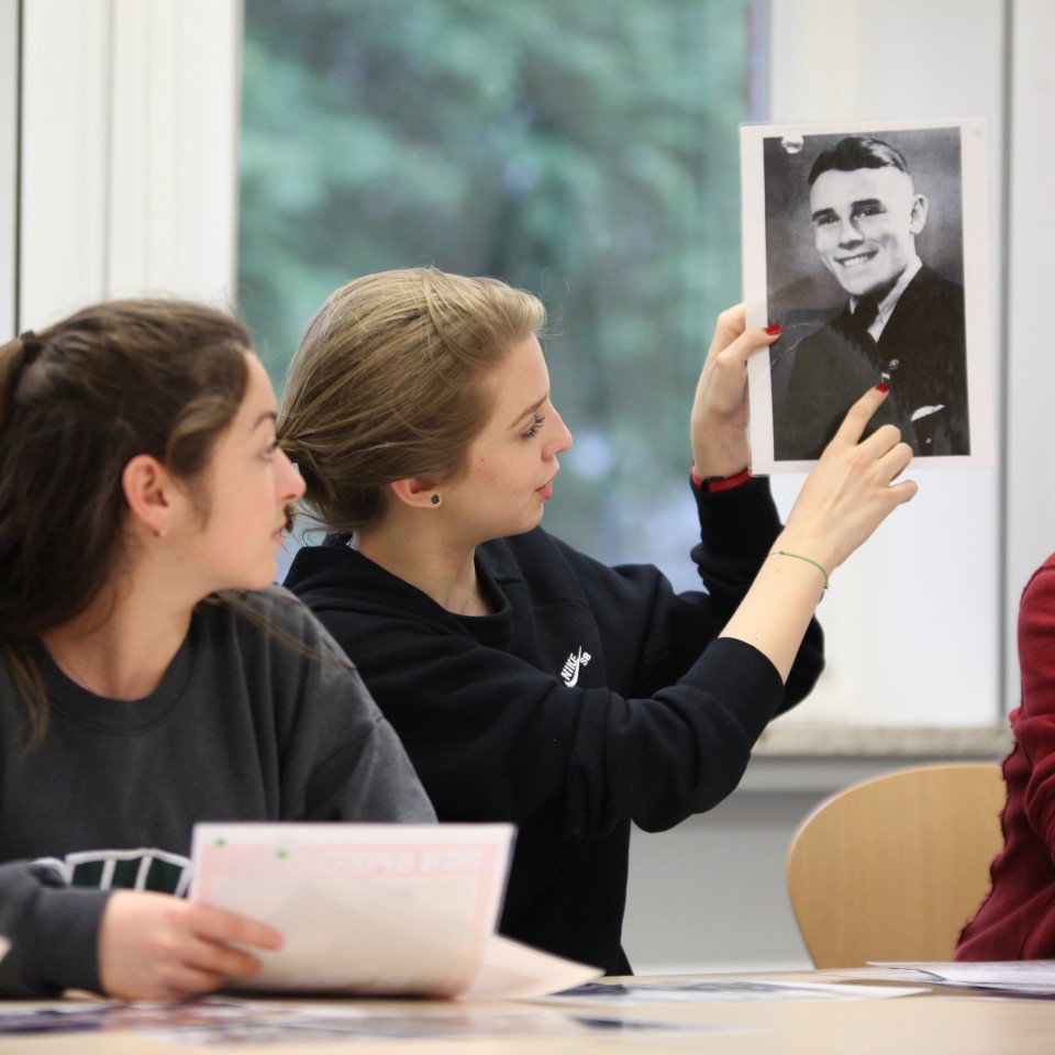 In einem Seminar zeigt eine Schülerin auf ein historisches Porträtfoto, auf dem ein lächelnder Mann zu sehen ist.