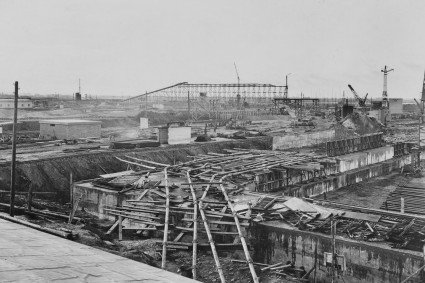 Schwarz-Weiss-Foto einer menschenleeren Großbaustelle. Es sind Kräne und Baumaterialien abgebildet. 