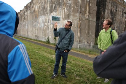 Ein freier Mitarbeiter steht mit seiner Gruppe am Deich am Denkort Bunker Valentin und präsentiert ein Foto des Bunkers.