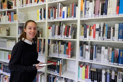 Eine lächelnde Praktikantin steht vor einem Bücherregal. Sie hält ein Buch in ihren Händen.