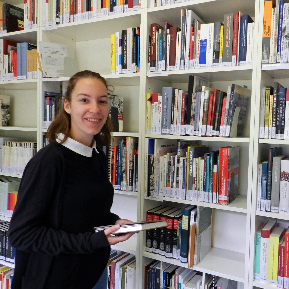 Eine lächelnde Praktikantin steht vor einem Bücherregal. Sie hält ein Buch in ihren Händen.